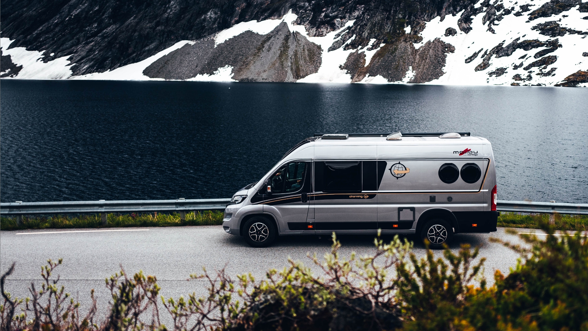 Ein Malibu Wohnmobil fährt auf einer Straße, mit Leitplanke, am Ufer eines Sees mit schneebedeckten Bergen im Hintergrund entlang